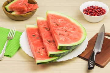 Delicious watermelon on a light wooden background. Three slices of berries ripe red watermelon on a white plate standing on the table with fork, knife. Green napkin, cranberries