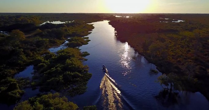Camera follows the boat sailing on the river towards the sunset. Aerial image of meander in the Pantanal Biome. Vegetation of Native forest. Top view. Mato Grosso do Sul state, Central-Western Brazil.