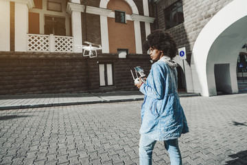 Pensive young biracial curly lady is driving white modern drone using remote controller having screen for showing video from quadrupter's camera while standing on pavement of urban street on sunny day