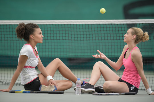 Women sat by tennis court net throwing ball into the air