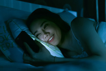 Asian woman on bed late at night texting using mobile phone tired falling sleep.