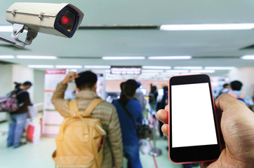 hand using smart phone monitoring and CCTV security indoor camera system operating in airport with people queue at immigration control background, surveillance security and safety technology concept