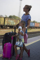 Mother and small daughter standing on railway station platform. With suitcases and a doll.