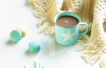 A blue cup with cocoa and macaroons, a warm plaid and notebooks for text on a white wooden background.. Copy space. Flat lay