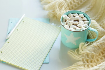 A blue cup with cocoa and marshmallows, a warm plaid and notebooks for text on a white wooden background.Top view. Copy space. Flat lay