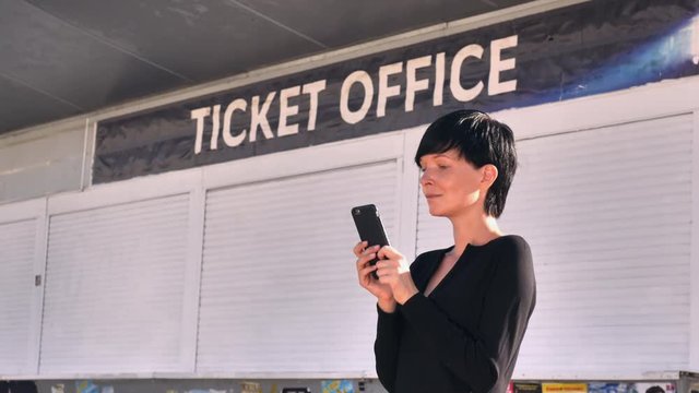 Businesswoman standing on the street near signboard ticket office using smart phone. Adult woman messaging or using application on mobile in city. Brunette with short hair wearing black dress on the