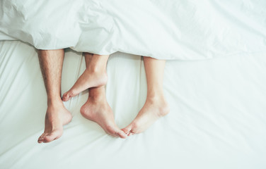 The feet or foot of couples sleeping on the bed and cover by the white blanket on a bright Sunday morning.