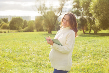 Fototapeta na wymiar Pregnant woman walking in park sunny summer outdoor mother touching tummy writing diary notebook