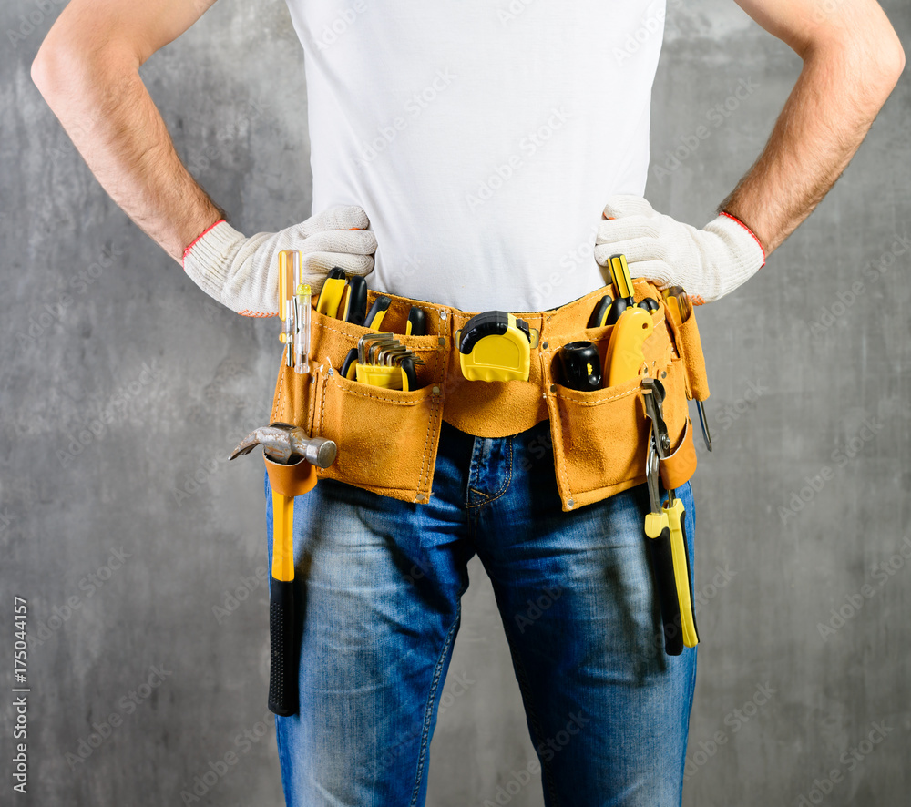 Wall mural unknown handyman with hands on waist and tool belt with construction tools against grey background. 