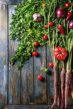 Raw organic purple carrot with variety of vegetables tomatoes, pepper, chard, garlic, onion on haulm over old wooden plank background. Top view with space. Food background.