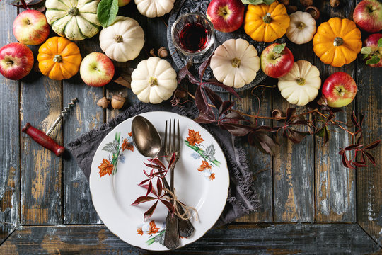 Autumn holiday table decoration setting with decorative pumpkins, apples, red leaves, empty plate with vintage cutlery, red wine, candle over wooden table. Rustic style. Flat lay
