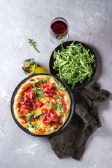Whole homemade pizza with cheese and bresaola, served on black plate with fresh arugula, olive oil, glass of red wine and textile napkin over gray concrete texture background. Flat lay.