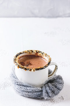 Vintage mug in wool scarf of hot chocolate, decor with nuts, caramel, spices over white texture background with space.