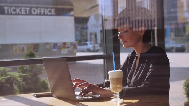 Professional businesswoman sitting in cafe typing on computer. Caucasian brunette enjoy morning and coffee checking email or chatting in social medial or working with documents sitting at the table in