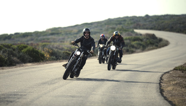 Three men wearing open face crash helmets and sunglasses riding cafe racer motorcycles along rural road.