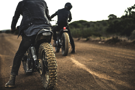 Rear view of two men sitting on cafe racer motorcycles on a dusty dirt road.