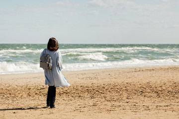 Young woman (brunette) in a light blue cardigan and navy jeans, with a striped backpack, walks along the beach.