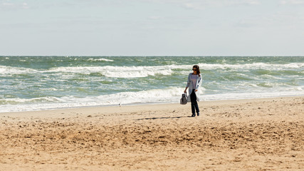 Young woman (brunette) in a light blue cardigan and navy jeans, with a striped backpack, walks along the beach.