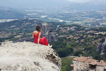 Naklejka premium Alone girl in a red dress on the edge of the rock at the monasteries of Meteora. Female on the rock and monasteries of Meteora in Greece in Thessaly