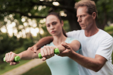 Girl doing exercises with dumbbells in the park. A man helps her. They are smiling