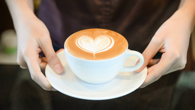 woman serving coffee while standing in coffee shop. Focus on Latte art hearth shape cup in female hands while placing of coffee on counter