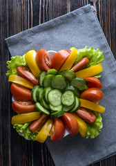 Sliced vegetables. Cucumbers, peppers, red and yellow tomatoes, lettuce on a plate on a wooden background. Gray napkin. Vertical shot.