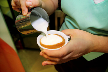 Barista making cappuccino in his coffee shop or cafe. Barista pouring latte froth to make a coffee latte art into the white coffee cup. Female hand pours milk into a bowl full of coffee. Hot drink.