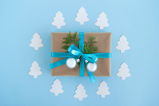 Gift box wrapped of craft paper, blue ribbon and decorated fir branches and silver Christmas balls on the blue background with paper fir tree, top view. Christmas present.