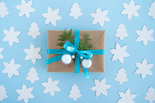 Gift box wrapped of craft paper, blue ribbon and decorated fir branches and silver Christmas balls on the blue background with white paper fir tree and star, top view. Christmas present.