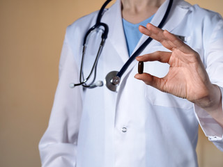 A female doctor with a stethoscope on a beige background holds a dark brown pill.