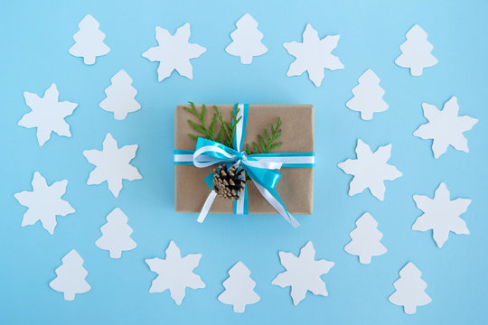 Gift box wrapped of craft paper, blue and white ribbons and decorated fir branches and pinecone on the blue background with paper white fir tree and star, top view. Christmas present.