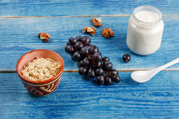 Healthy breakfast ingredients.Homemade yogurt in glass jars, ceramic bowl with oat flakes, walnuts, grapes on light blue background, copy space