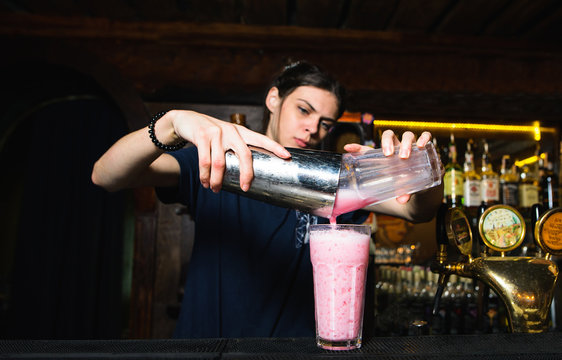 The girl bartender creates a beautiful, pink, alcoholic cocktail. The barman pours the ingredients from the shaker into a glass