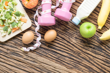 Healthy eating, dieting, slimming and weight loss concept - Top view of green apple, bananas, measuring tape, dumbbells, eggs, milk bottle and vegetables block kerry and carrot on wooden background. 