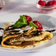 Serving of homemade crepes with strawberries, chocolate topping and powdered sugar. Small jars of red berries and teacup in background.