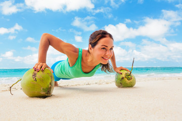 Active woman doing fitness exercise plank on coconut to keep fit and health. Beach surf background. Healthy lifestyle, morning workout, sport activity on summer family vacation in tropical island.