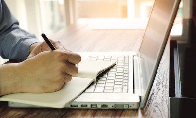 a man is writing some word on notebook with laptop in office room