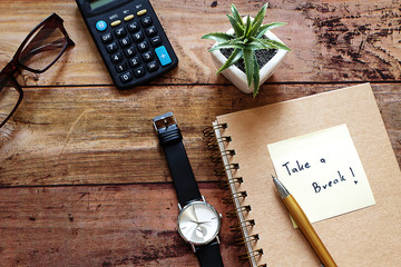 Wood Desktop work space have brown glasses,calculator,pot plant ,black watch,pen,notebook,and note...