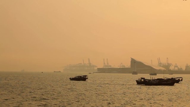 Traditional Chinese fishing boats sailing at sea with Shenzhen west port with container ship and cranes in background, back-lit by sunset light; Guangdong province, People's republic of China; 