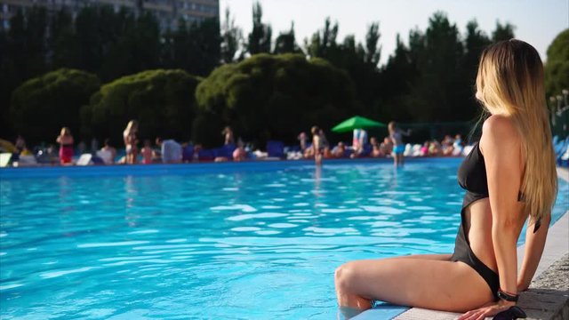 Cute young woman is sitting by the pool and getting her legs wet. She is taking sunbath and enjoying water.