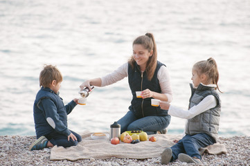 smiling beautiful mother filling kids cups with hot tea sitting by the sea in winter