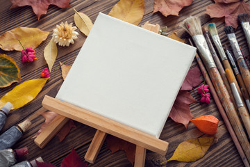 Canvas on easel, paint tubes, brushes for painting and autumn leaves on old wooden desk. Top view. Flat lay. Retro toned.