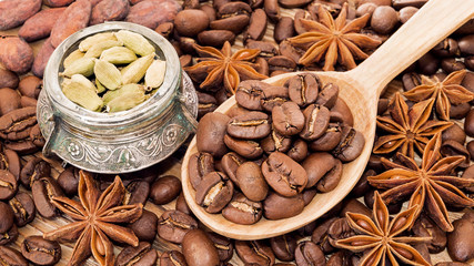 Coffee beans in a wooden spoon with cardamom, cocoa beans and anise illicium as warm aromatic background