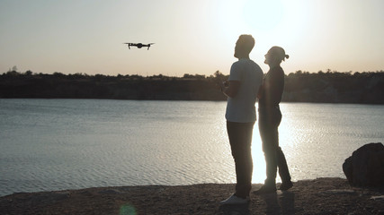 Silhouettes of two men on river bank using remote controller and quadcopter in sunlight.