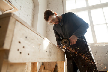 Worker in protective eyewear working in small workshop interior using die angle grinder for cutting steel bar, throwing sparks from metal grinding machine with abrasive disk wheel, making furniture