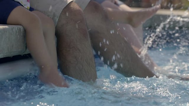 Mom, dad and infant daughters sit on side of outdoor swimming pool splashing their feet in the water. Detail side view, slow motion 4K recorded at 60fps.