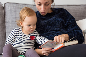 Eighteen months old toddler girl sitting by her mother's side, reading the book; concept of education and parenting and early age development