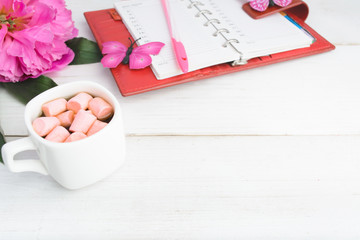Coffee with marshmallow with planning notebook and flower over white wooden background