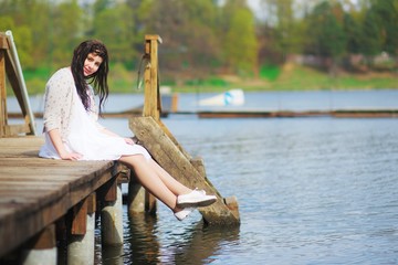 Portrait of young pretty woman brunette in a white dress with flowing long hair, sitting on a wooden bridge, lowering his feet over water, in the bright sunshine