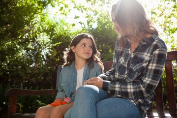 Girl sitting with mother on wooden bench against trees during